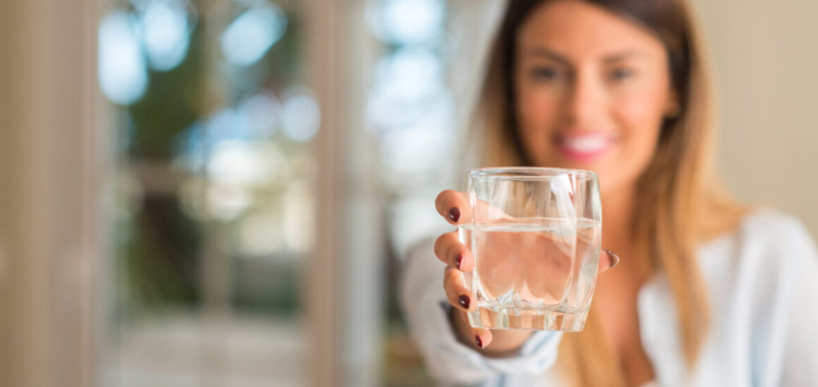 Beautiful young woman smiling while holding a glass of water at home. Lifestyle concept.