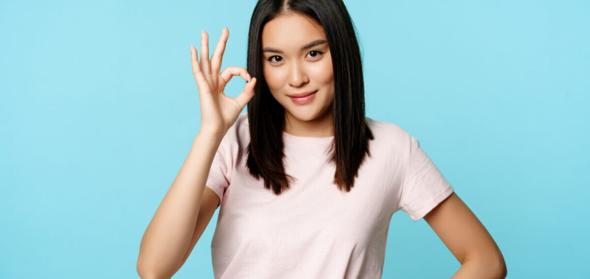 Smiling asian woman showing okay sign, gives approval, recommends smth good, standing over blue background