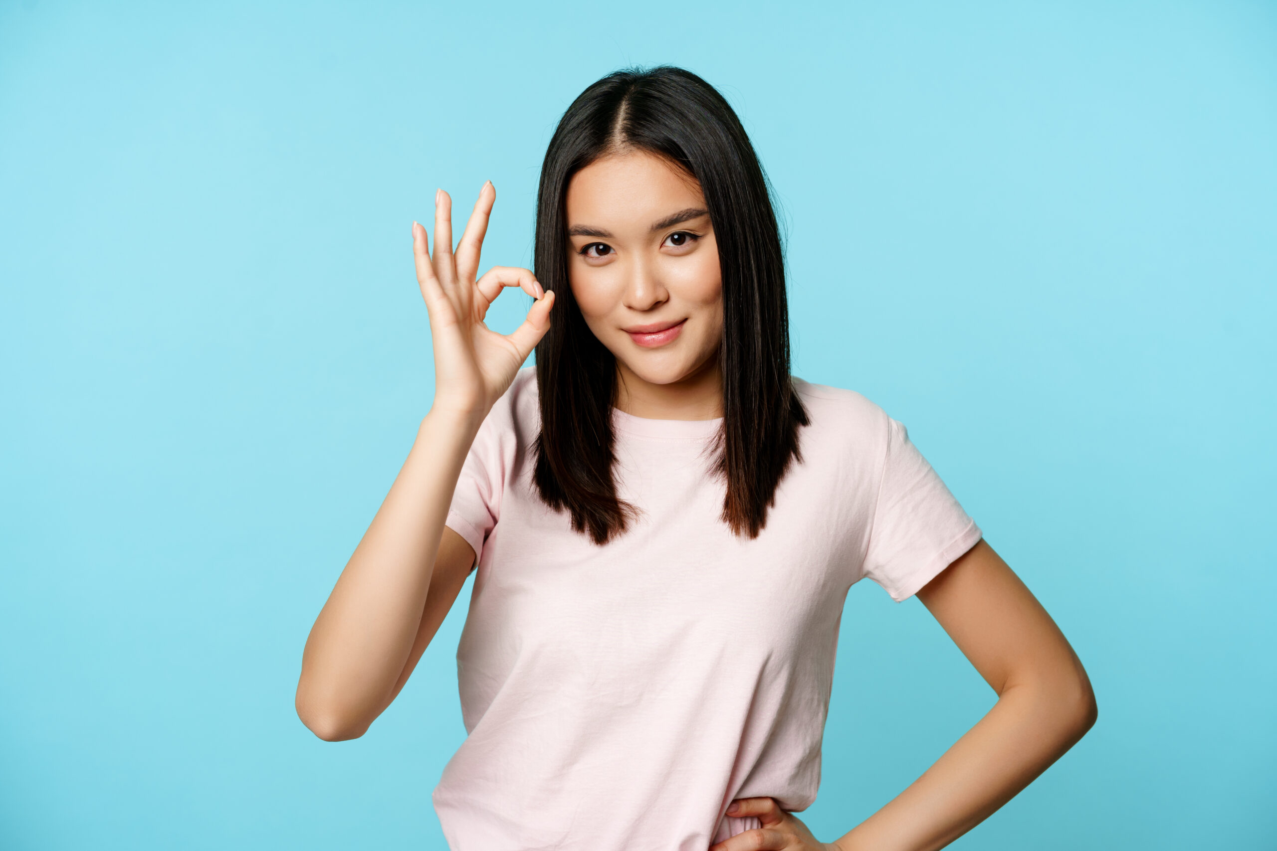 smiling asian woman showing okay sign gives approval recommends smth good standing blue background scaled